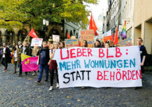 Mehrere Menschen demonstrieren für mehr Wohnungen. Sie halten Fahnen und Plakate in den Händen.