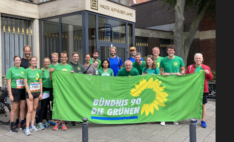 Eine Gruppe von Menschen hat vor sich ein Banner aufgestellt. Das Banner hat einen grünen Hintergrund, auf dem das Logo der Partei BÜNDNIS 90/DIE GRÜNEN zu sehen ist: Der Name Die GRÜNEN in weißer Schrift, dazu eine gelbe Sonnenblume.