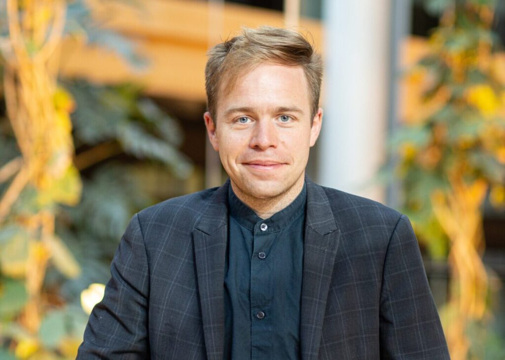 Portrait eines weißen Mannes mit blonden Haaren, von dem man nur den Oberkörper sieht. Er trägt einen dunkelgrauen Anzug und ein dunkelblaues Hemd.